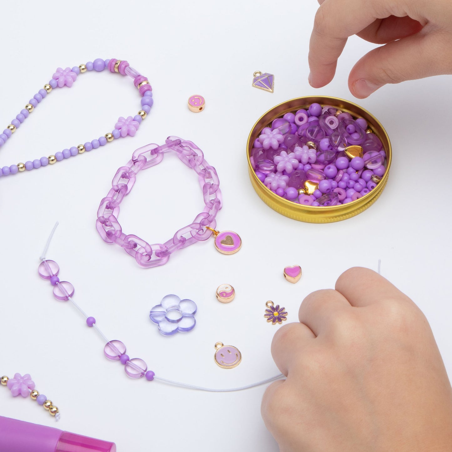 Charms and bracelets on a table top. The tube lid is filled with beads and a child is choosing a bead to string onto the bracelet she is holding.