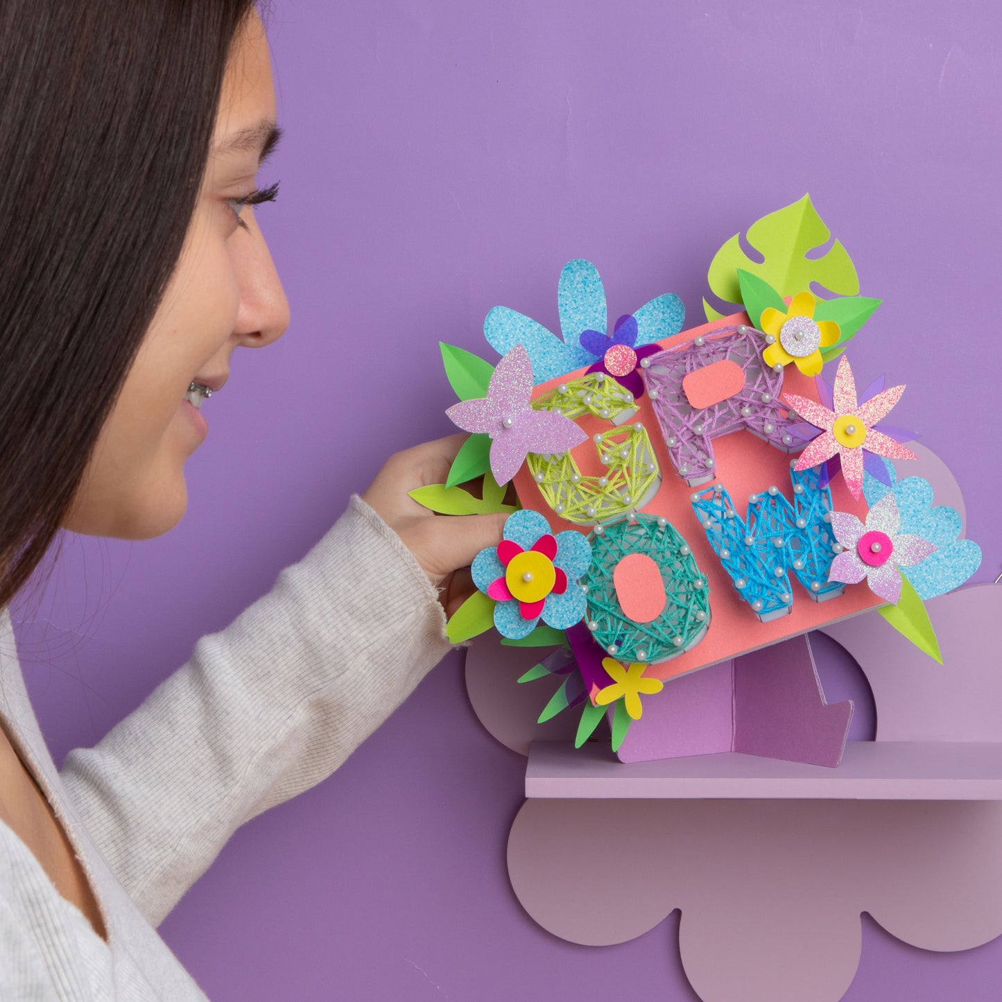 "GROW" string art being placed on a flower shelf with a smiling girl. The background is purple.