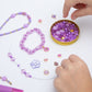 Charms and bracelets on a table top. The tube lid is filled with beads and a child is choosing a bead to string onto the bracelet she is holding.